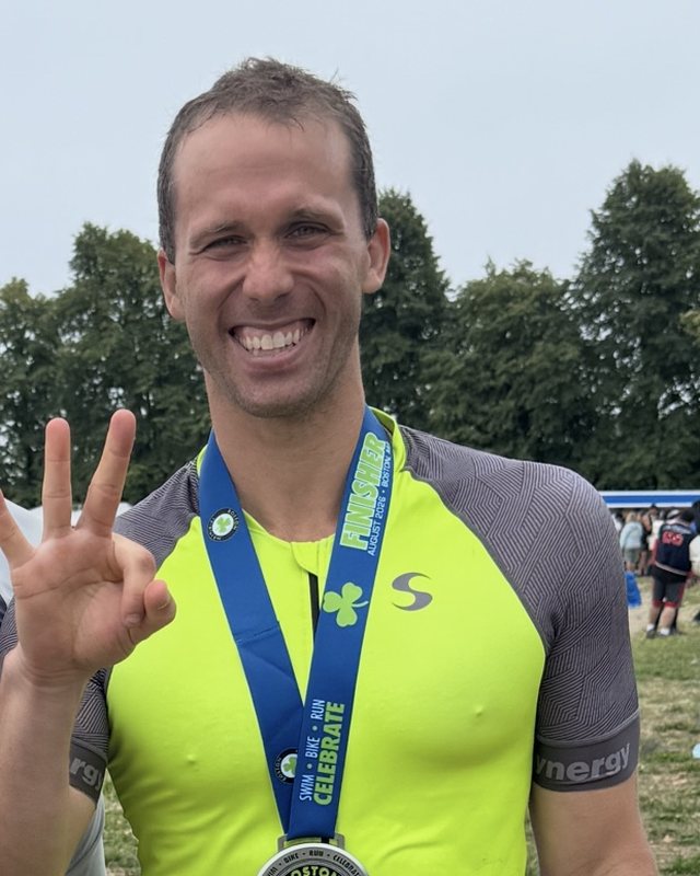 Brooks Robinson smiling at the Boston Triathlon finish in a yellow-green tri suit, wearing the finisher medal