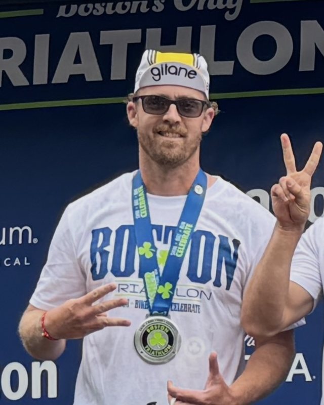 Ian Swett smiling at the Boston Triathlon, wearing a white Boston Triathlon t-shirt and the finisher medal